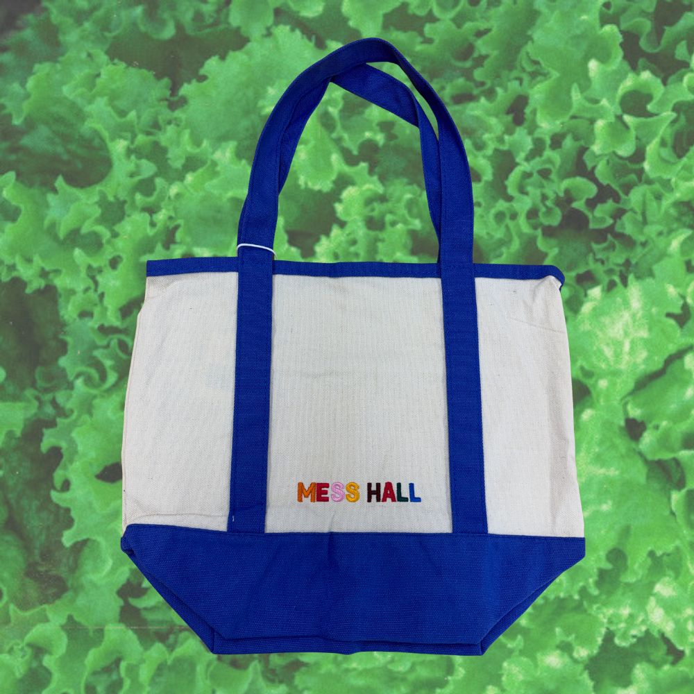 A white and blue canvas tote bag with 'MESS HALL' printed in colorful letters on the front, set against a green textured background.