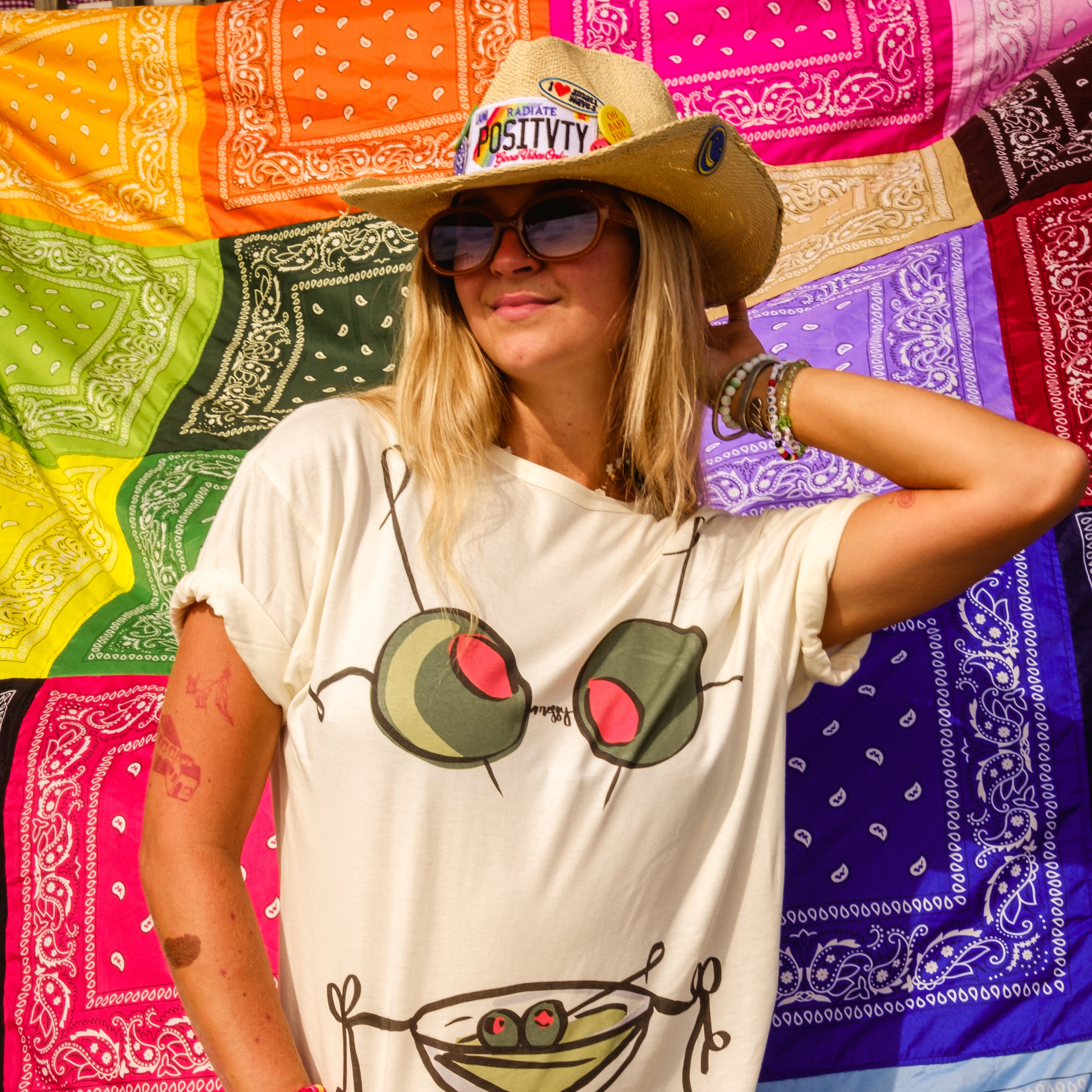 A woman wearing a cream-colored graphic t-shirt with an olive-themed design, a straw cowboy hat, and sunglasses, posing in front of a colorful rainbow bandana backdrop.