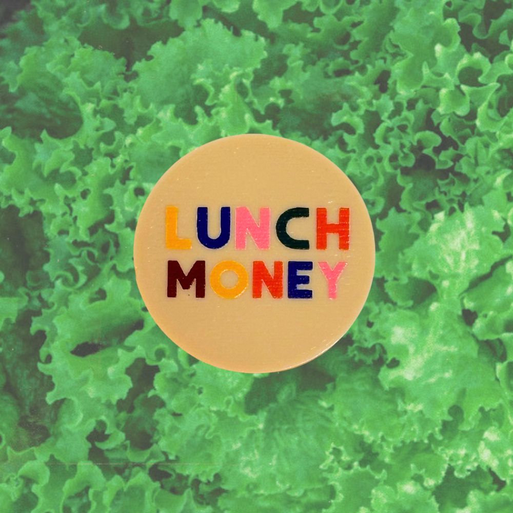 A round beige enamel pin featuring the text 'LUNCH MONEY' in colorful bubble letters, set against a background of green lettuce leaves.
