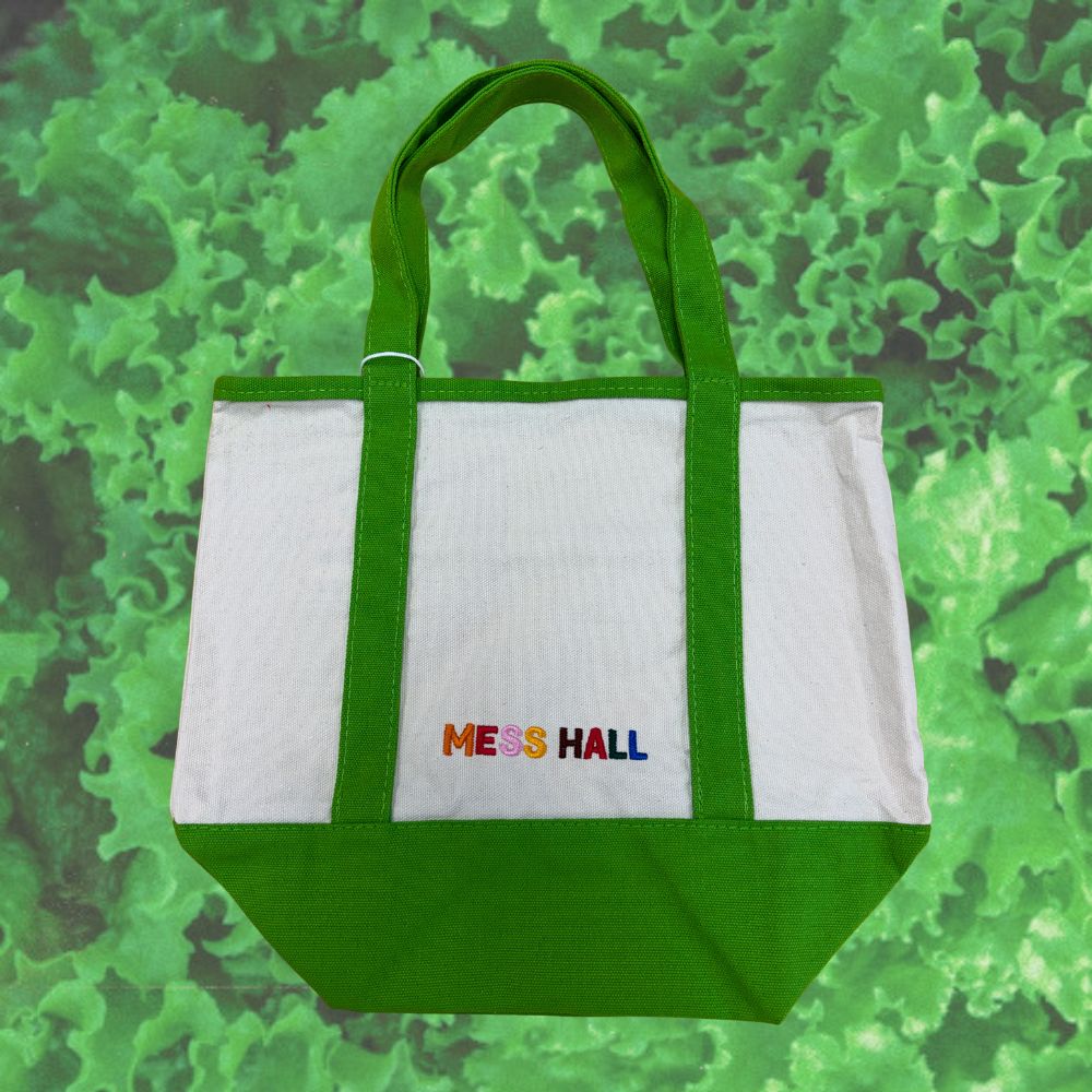 A white and green canvas tote bag with 'MESS HALL' embroidered in multicolor letters on the front, set against a green textured background.
