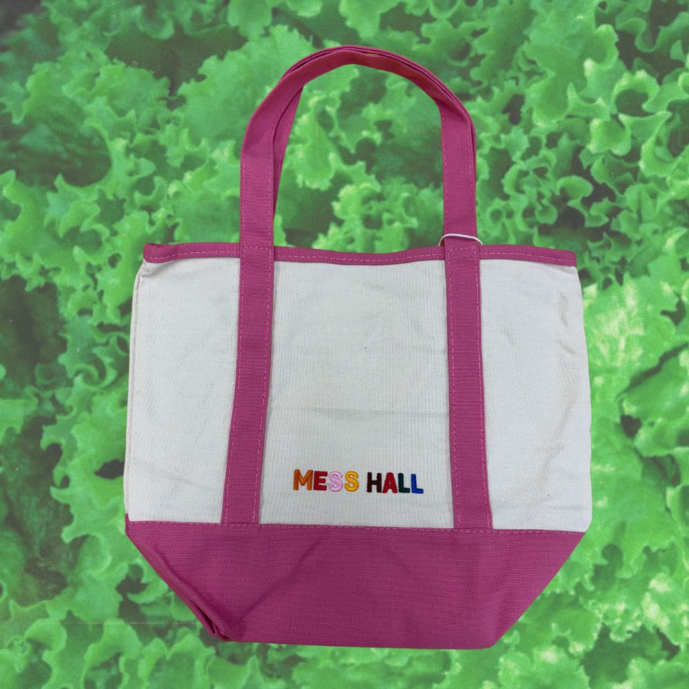 A white canvas tote bag with pink handles and bottom trim, featuring the brand text 'MESS HALL' embroidered in multicolored letters on the front, set against a green leafy background.