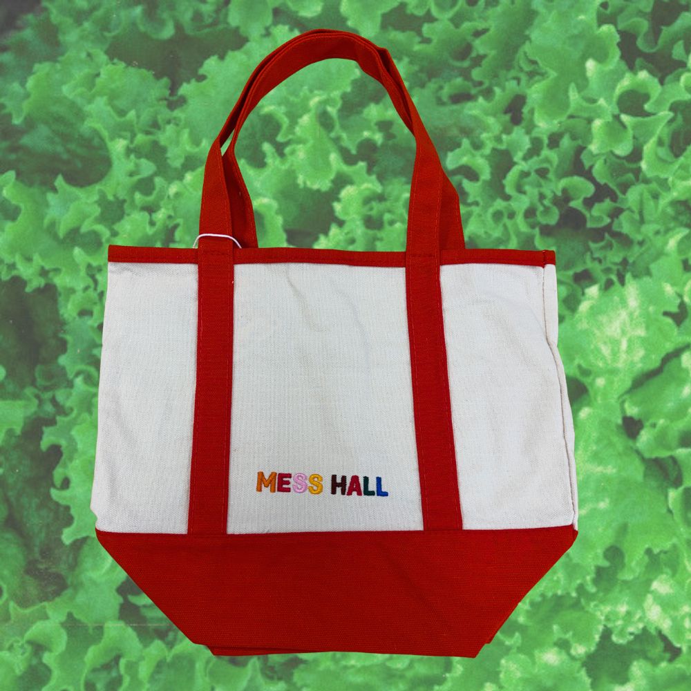 A white canvas tote bag with red handles and a red bottom panel, featuring the text 'MESS HALL' embroidered in multicolor letters on the front, set against a green textured background.