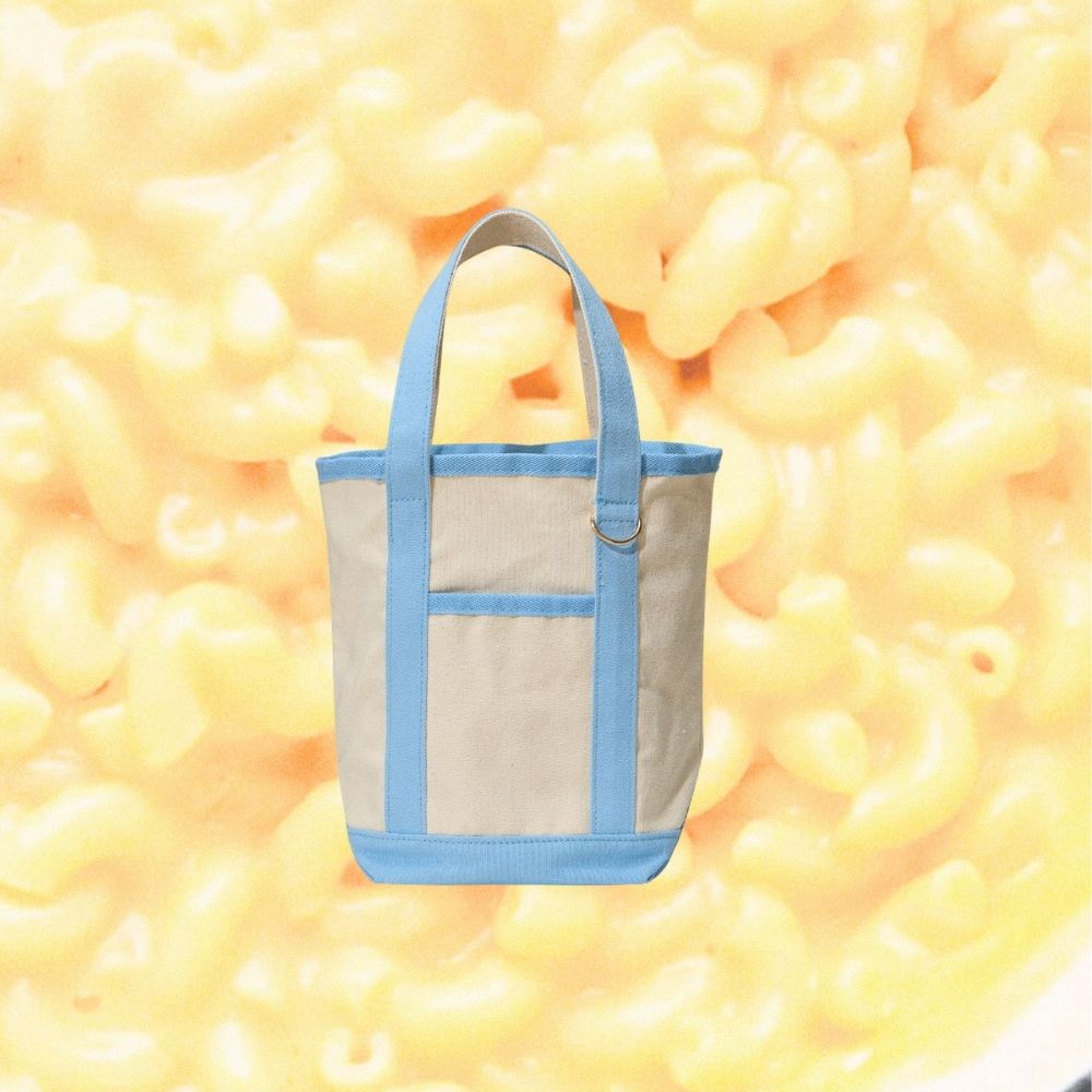 A canvas tote bag with light blue trim and handles, featuring a front pocket, centered against a background of yellow macaroni pasta.