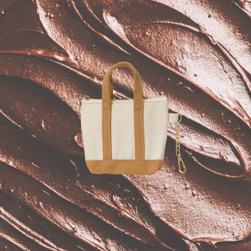 A small canvas tote bag keychain with cream white panels and tan leather-like trim, handles, and base, shown against a glossy brown chocolate-like textured background. A short gold chain is attached on the right side.