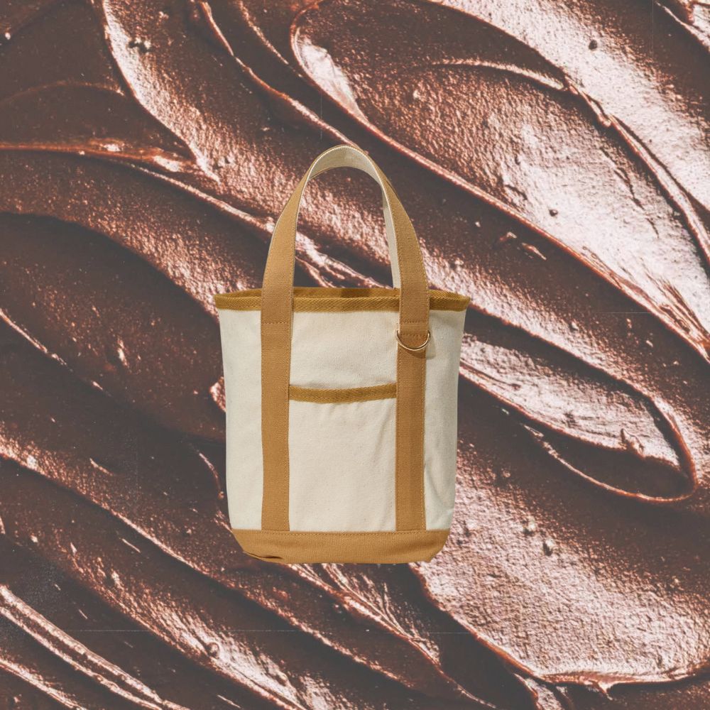 A cream canvas tote bag with tan trim and handles, centered against a textured background of dark brown chocolate frosting swirls.