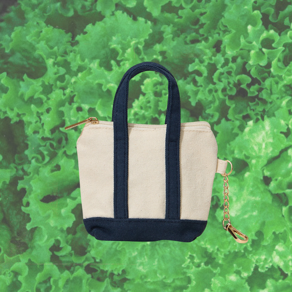 Small tote bag keychain with navy handles and beige base on a green leafy background