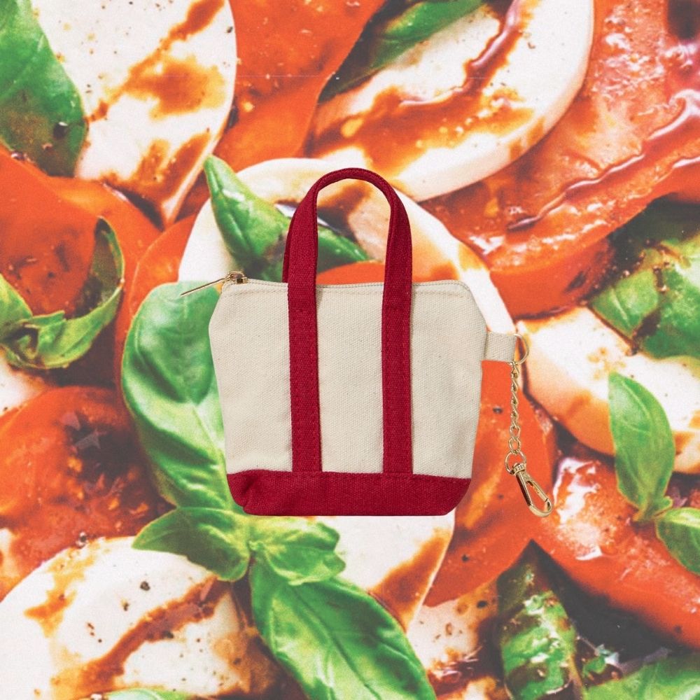 A small canvas tote bag with cream panels and dark red trim, including the handles and bottom band, placed over a colorful food background of tomato, mozzarella, and basil.