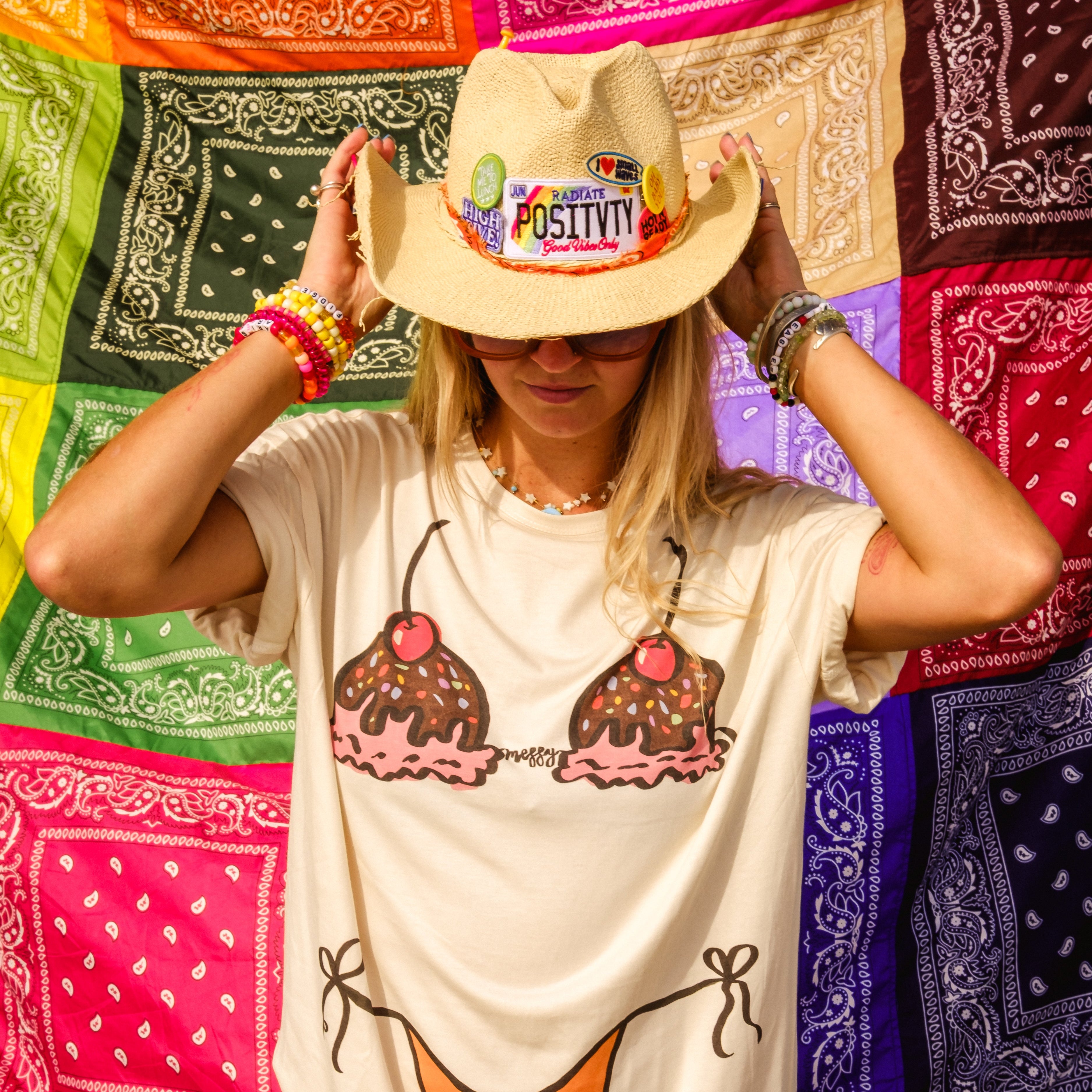 A woman wearing a cream-colored graphic t-shirt dress with an ice cream cone design, a straw cowboy hat with a 'Radiate Positivity' patch, and colorful bracelets, posing in front of a colorful bandana-patterned backdrop.