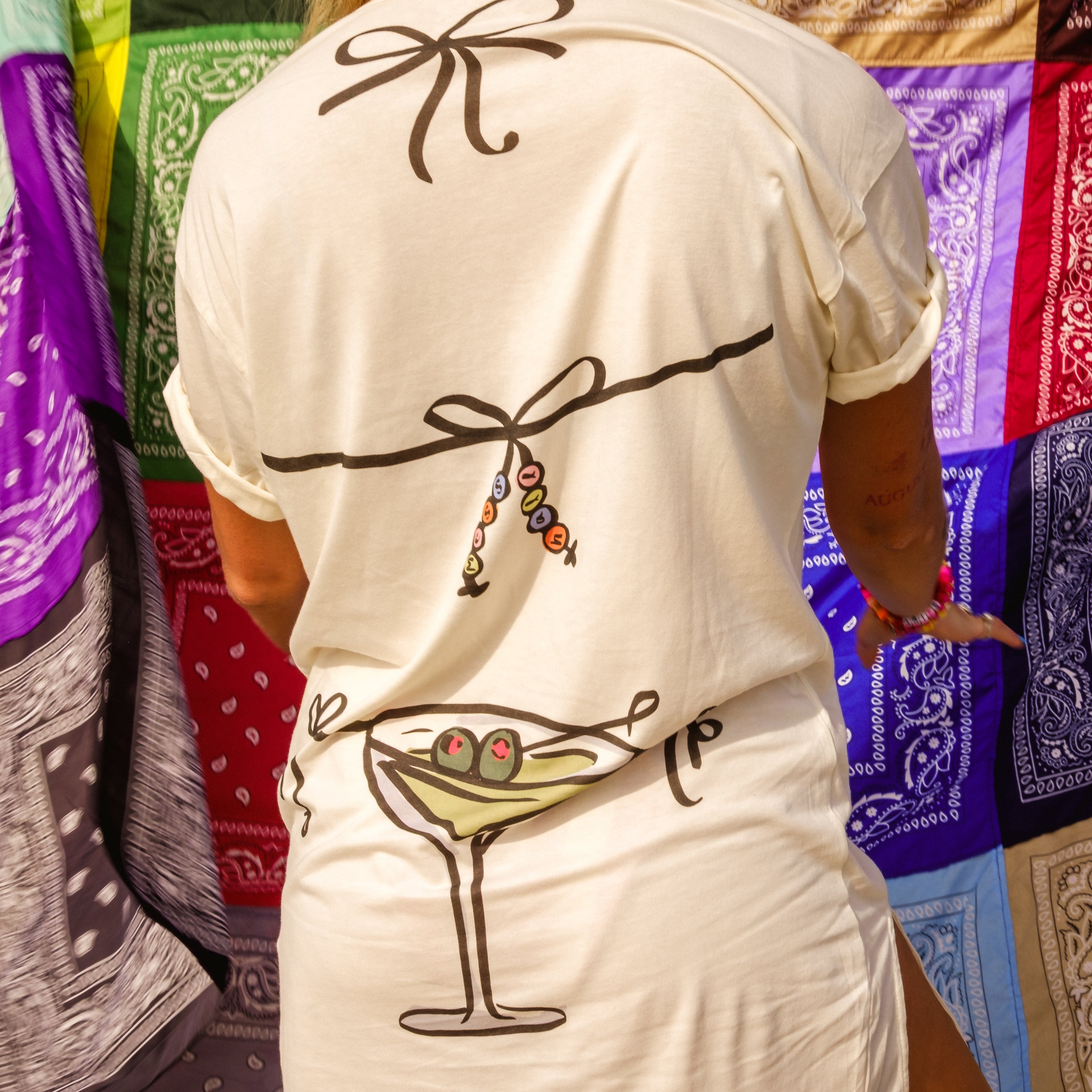 A woman shown from behind wearing a cream-colored t-shirt dress with black line-art illustrations of bows and a martini glass with olives, set against a colorful background of bandanas.